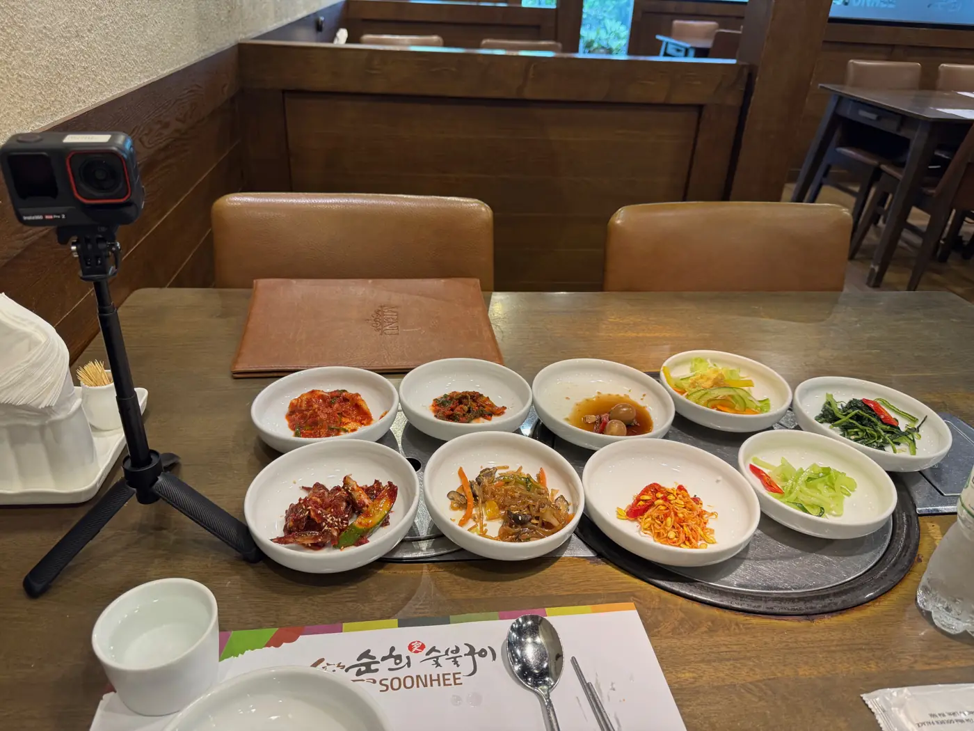 식탁에 깔린 반찬 아홉 가지와 순희 숯불구이 종이 깔개, 옆에 세운 카메라